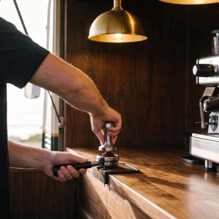 A barista's hands tamping ground coffee into a chrome portafilter.